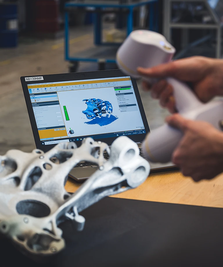 Eine Person scannt mit einem tragbaren 3D-Scanner ein komplexes Metallobjekt, während das gescannte 3D-Modell auf einem Laptop-Bildschirm im Hintergrund angezeigt wird.