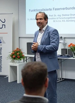 Ein Mann in einem hellblauen Anzug steht und spricht vor einem Publikum in einem Konferenzraum, während im Hintergrund eine Präsentationsfolie und Blumen zu sehen sind.