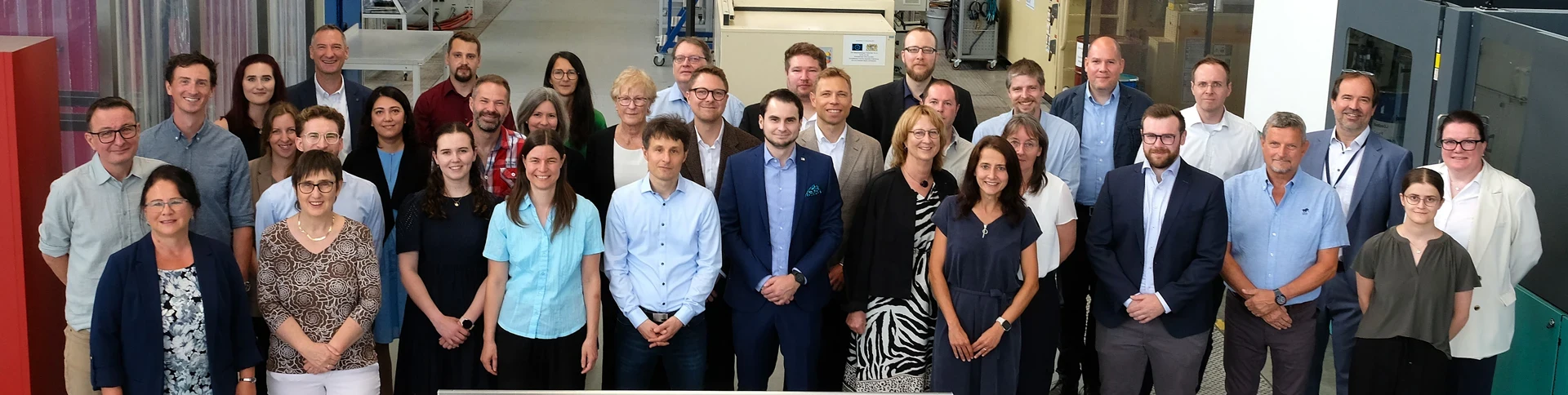 Eine Gruppe von etwa 36 Männern und Frauen in Geschäfts- und Freizeitkleidung steht in einem Innenraum dicht beieinander, lächelt und blickt in die Kamera für ein Gruppenfoto in einer Industrie- oder Büroumgebung.