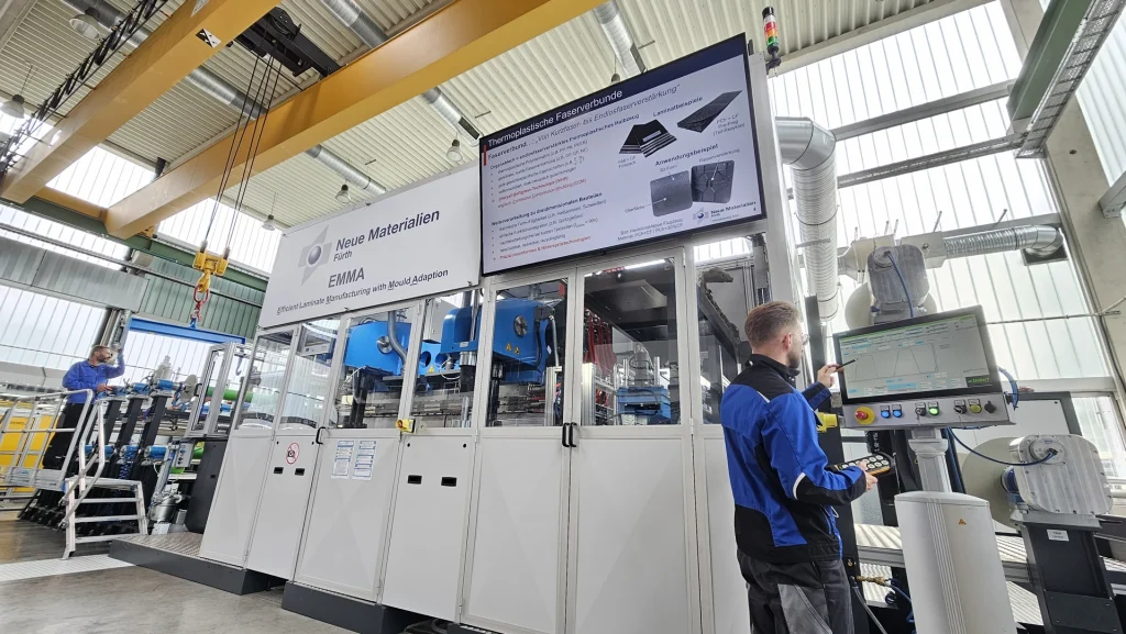 Ein Techniker bedient ein Bedienfeld neben einer großen Industriemaschine in einer hellen, modernen Fabrik. Über den Bildschirmen werden technische Informationen über neue Materialien und Produktionsverfahren angezeigt.