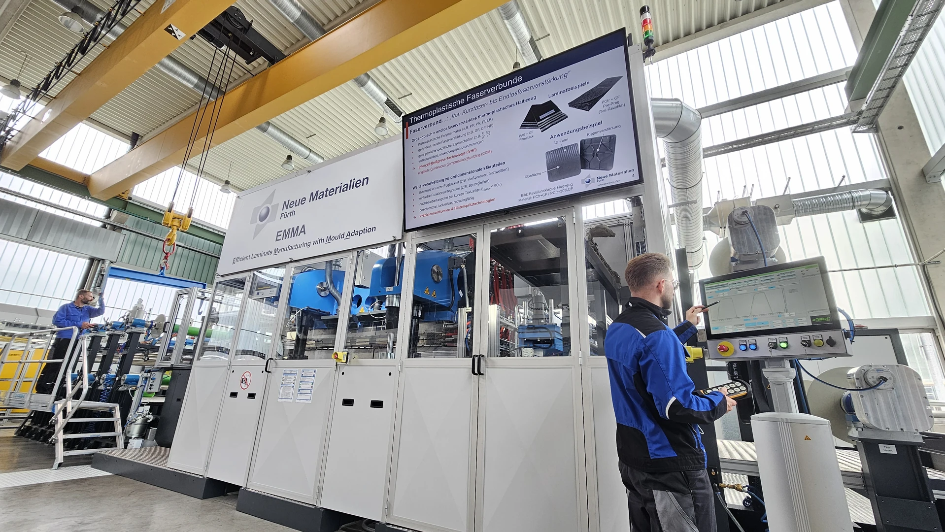 Ein Techniker bedient ein Bedienfeld neben einer großen Industriemaschine in einer hellen, modernen Fabrik. Über den Bildschirmen werden technische Informationen über neue Materialien und Produktionsverfahren angezeigt.