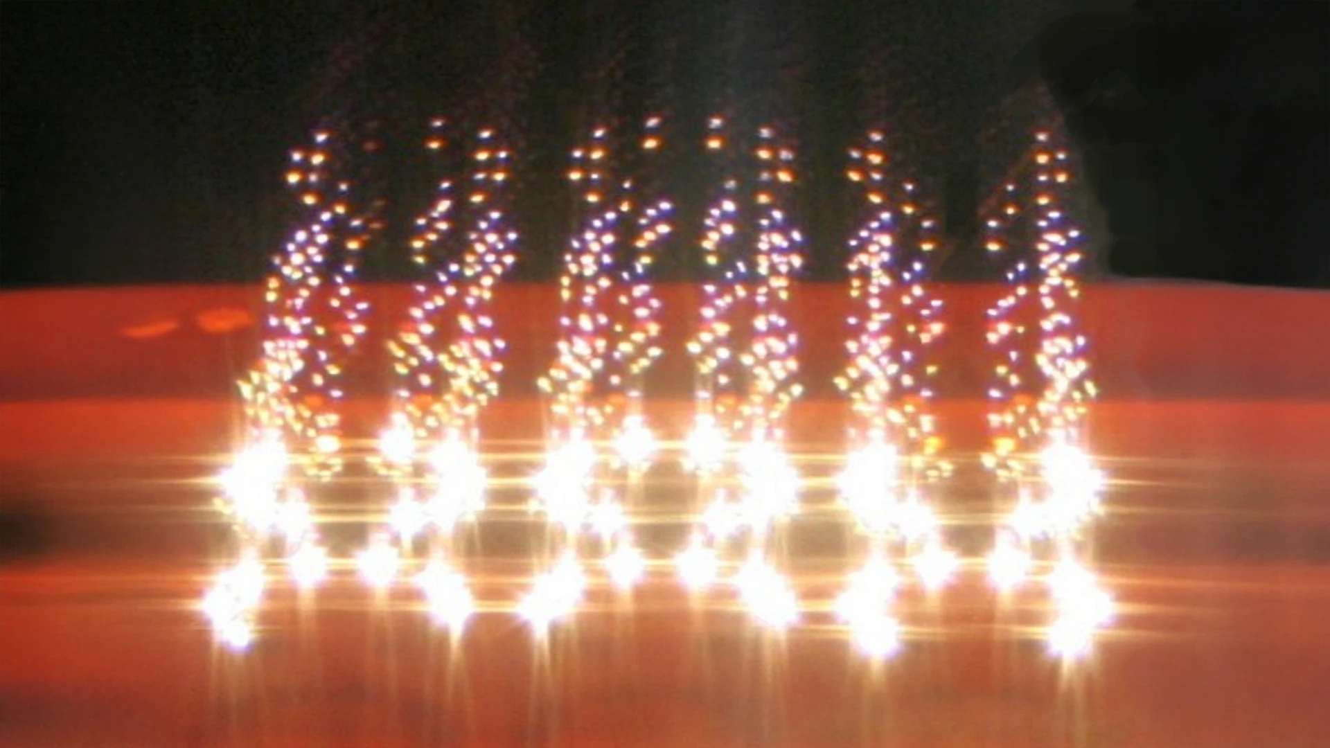 Reihen heller, weißer Lichter, die in einem Gittermuster angeordnet sind, werden von oben mehrfach auf einer glänzenden roten Oberfläche reflektiert, wodurch ein dynamischer Spiegeleffekt in einer dunklen Umgebung entsteht.