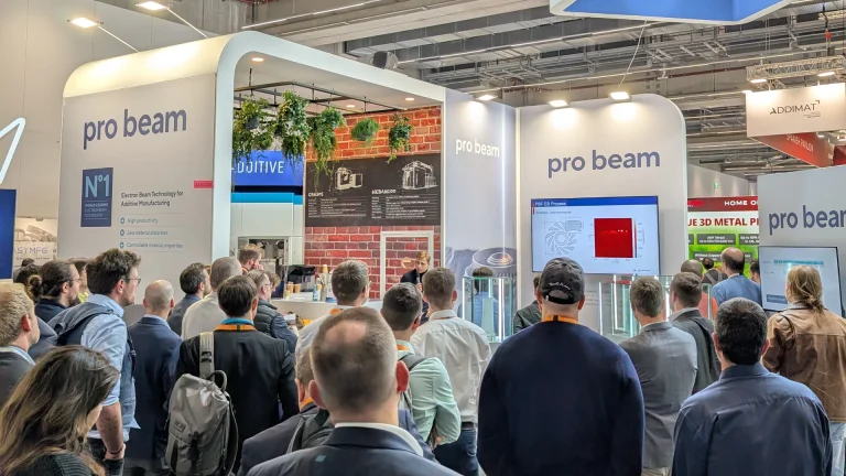 Eine Menschenmenge versammelt sich an einem Messestand von pro beam, der mit digitalen Bildschirmen, Informationsdisplays und moderner Dekoration mit Pflanzen ausgestattet ist. Die Besucher sehen sich eine Präsentation an und interagieren mit dem Exponat.