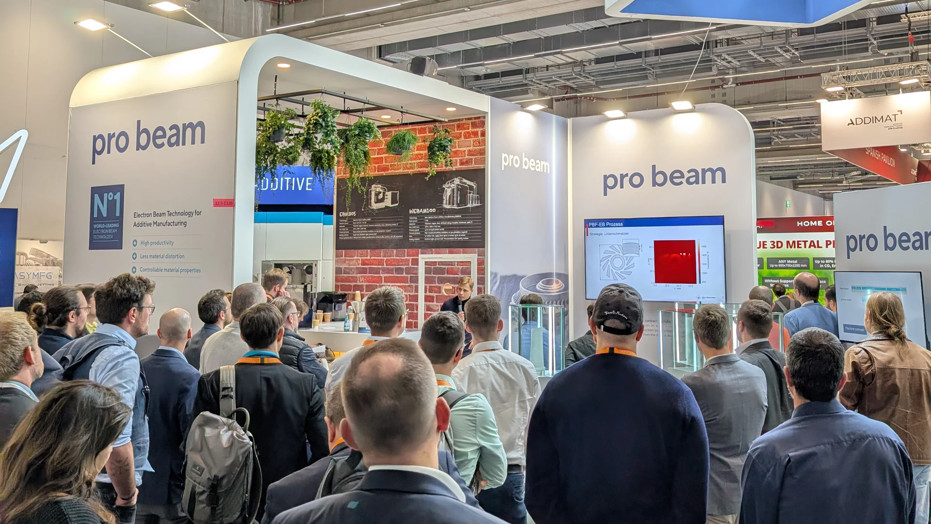 Eine Menschenmenge versammelt sich an einem Messestand von pro beam, der mit digitalen Bildschirmen, Informationsdisplays und moderner Dekoration mit Pflanzen ausgestattet ist. Die Besucher sehen sich eine Präsentation an und interagieren mit dem Exponat.