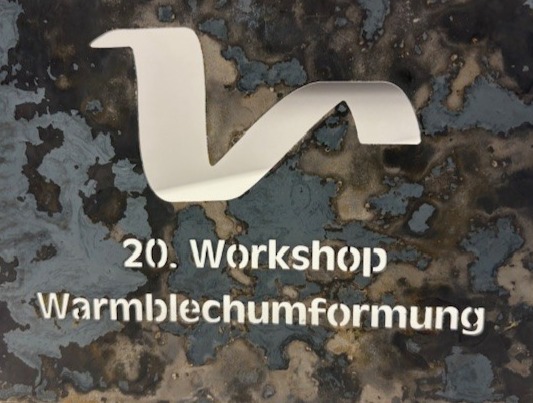 Ein Metallschild zeigt ein stilisiertes weißes Logo über dem Text 20. Werkstatt Warmblechumformung vor einem strukturierten, verrosteten Hintergrund.