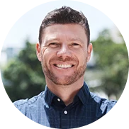 Ein Mann mit kurzen braunen Haaren und einem Bart lächelt. Er trägt ein blau kariertes Hemd und steht im Freien vor einem unscharfen Baum im Hintergrund.